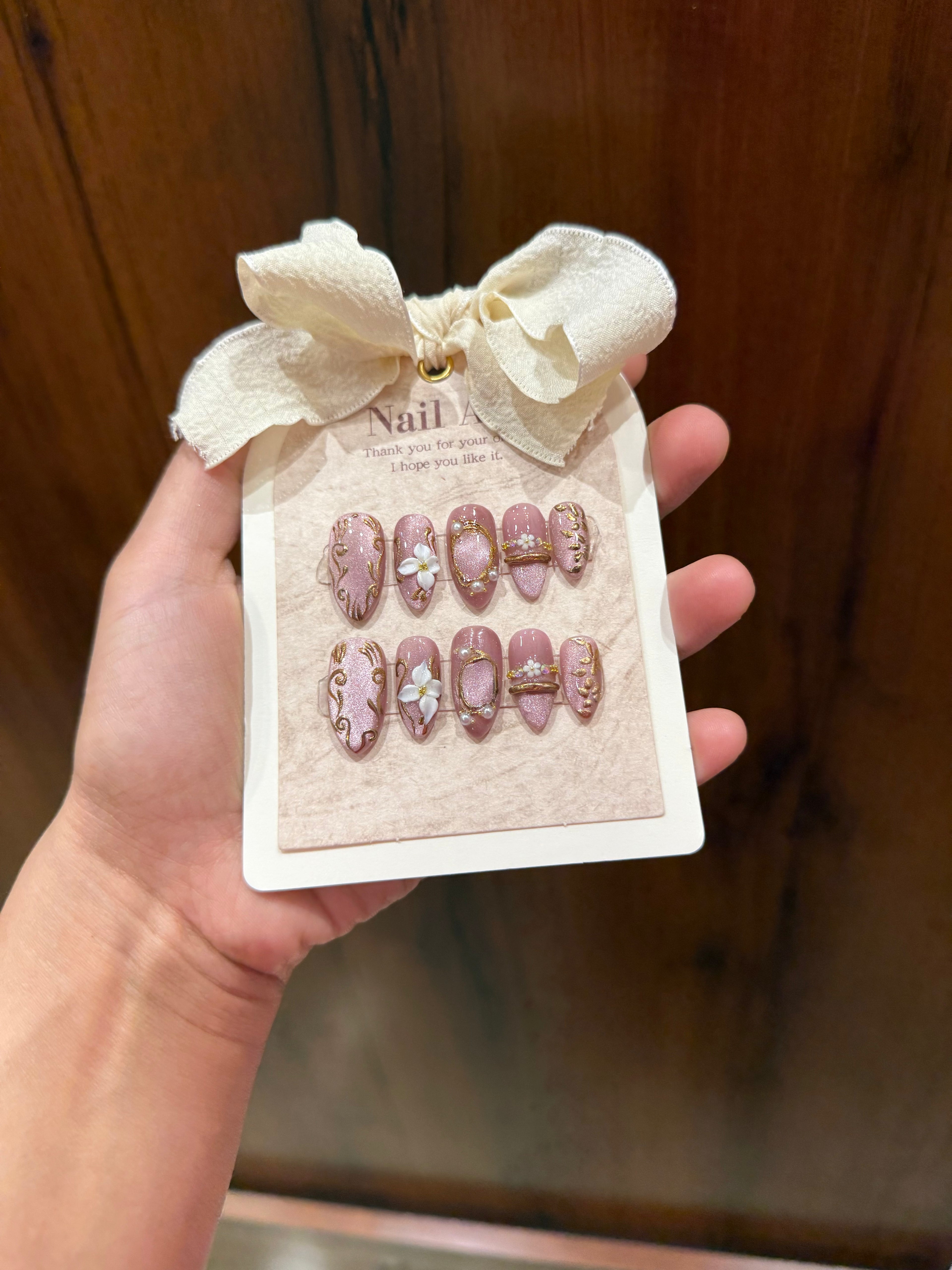 Set of decorative nail art extensions on a branded card held by a hand against a wooden background.